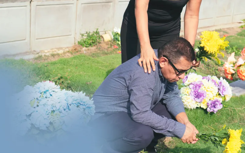 Hombre arrodillado junto a una tumba con flores mientras una mujer le toca el hombro