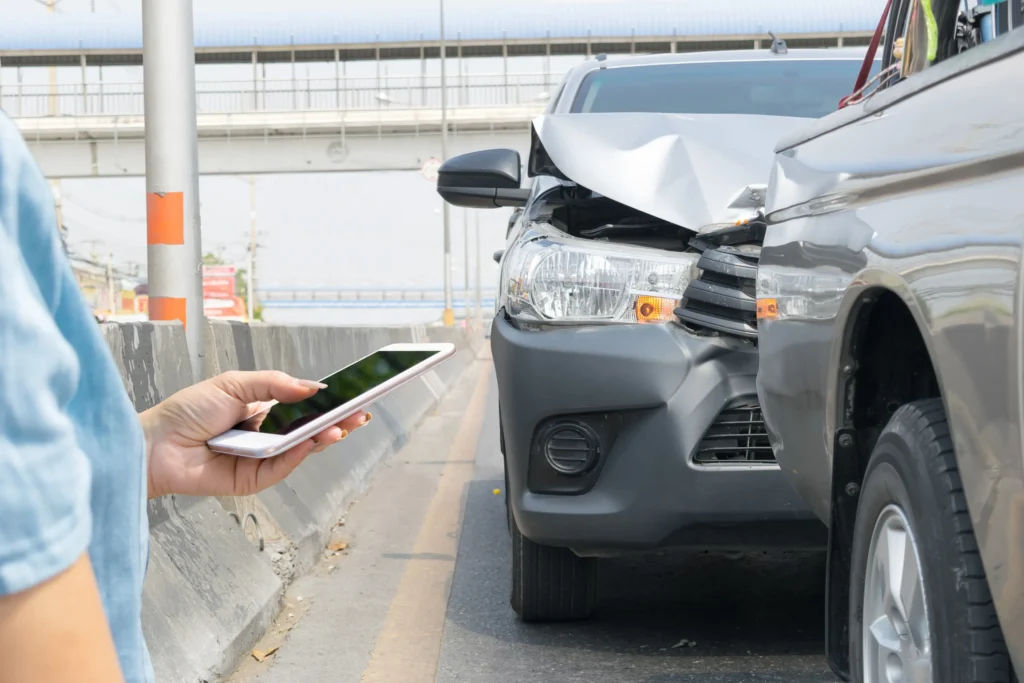 A person on their phone with a car crash accident behind them.